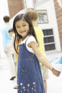 Photo of a young Asian preschooler, holding hands in a line of children heading into preschool.
