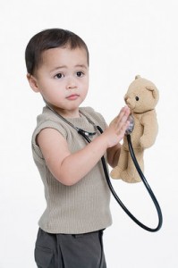 A young boy, holding the end of a stethoscope to his teddy bear's heart.