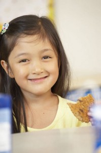 Photo of a first grader, holding a cookie and grinning. Foto de una estudiante del primer grado, sonriente, comiendo una galleta.