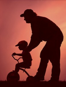 Silhouette image of a man steadying a youngster riding on a tricycle.