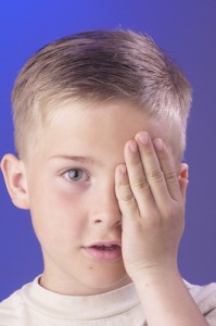 A young boy is taking an eye exam and covers one eye with his hand. Un muchacho toma un examen visual y está cubriendo un ojo con la mano.