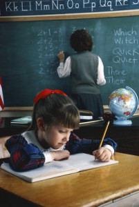 A student sits writing at her desk, while the teacher is in the background, writing on the chalkboard.