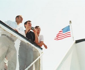 Photo of three men leaning on a railing looking out, and the U.S. flag flies nearby.