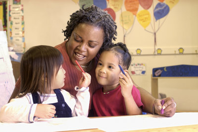 A preschool teacher has one arm around an Asian preschooler and the other arm around a grinning African-American preschooler.
