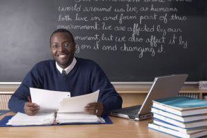 Teacher in front of class, stack of textbooks at his side and the chalkboard covered in writing