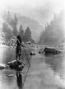 Native man stands alone atop a rock in a still river, fishing